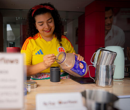 Preparing the perfect hot chocolate for our non-coffee drinking guests. A female barista wearing a yellow Colombia soccer jersey measuring chocolate powder into a cup.