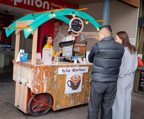 Bringing the café experience directly to your event with our fully equipped mobile setup. A wooden mobile coffee cart with a teal canopy serving two customers outdoors.